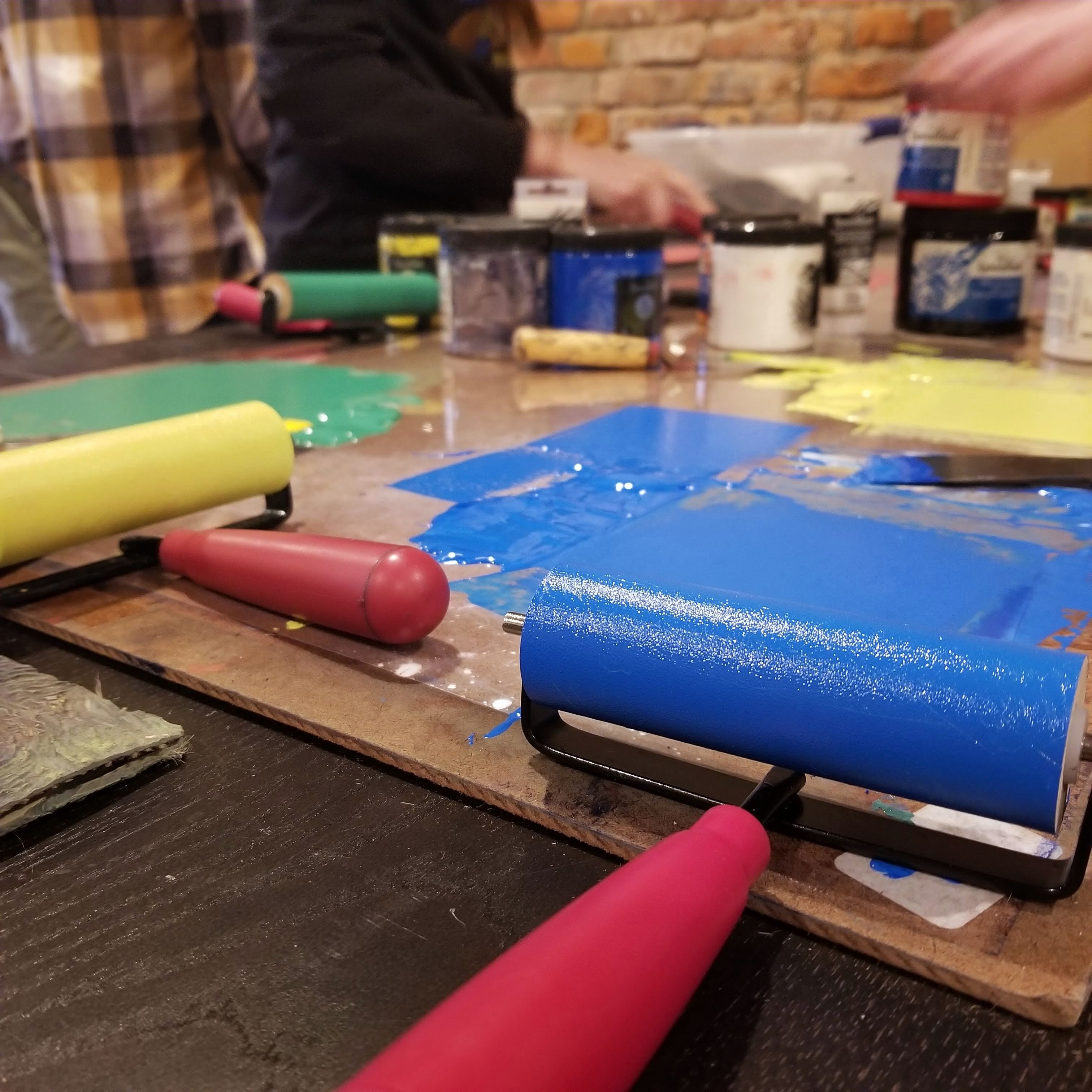 close-up photo of inked brayers on ink-mixing work surface, with people working in background; main ink colors and bright blue, yellow, and green