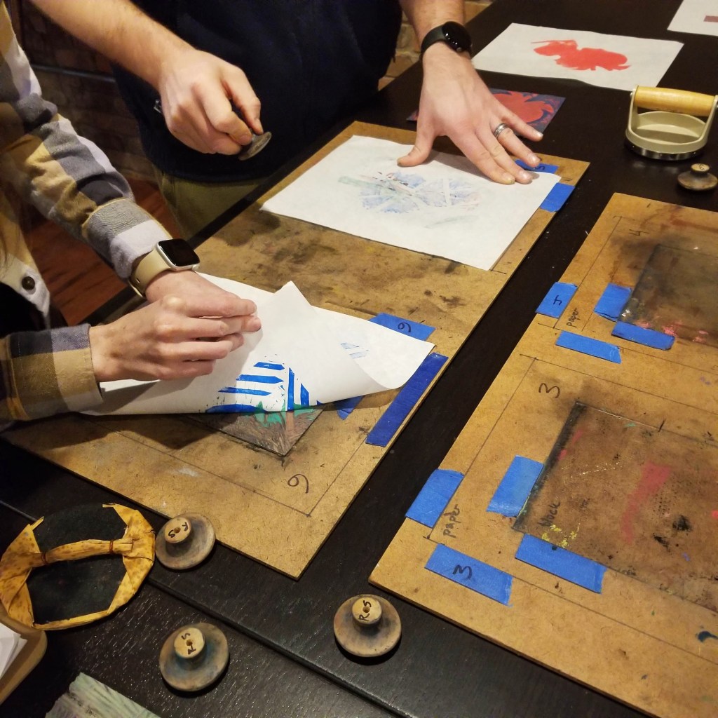 close-up photograph of two people hand-printing relief prints using a jig and applying pressure to the back of the paper; one person is peeling back one corner of their print to check progress
