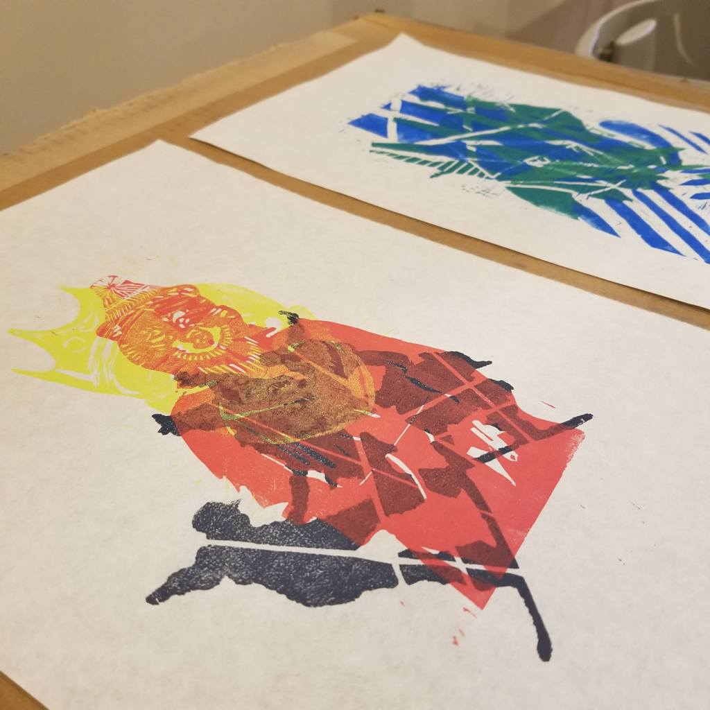 angled view of two layered, colorful relief monoprints; print in foreground contains imagery of snowboarders in silhouette and an anthropomorphized bear wearing a stocking cap and trenchcoat printed in pink; print in background is linear in blue and green ink